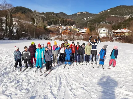 Eine Gruppe von Kindern und Erwachsenen posiert für ein Foto auf einem verschneiten Feld, mit Bergen und Häusern im Hintergrund.