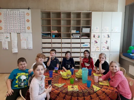 Eine Gruppe von Kindern sitzt an einem Tisch, möglicherweise in einem Klassenzimmer. Sie lächeln und scheinen für ein Foto zu posieren. Der Tisch ist mit einer bunten Tischdecke bedeckt. Auf dem Tisch stehen Becher und Schüsseln. Dahinter befinden sich Schränke und eine Pinnwand mit angehefteten Papieren.