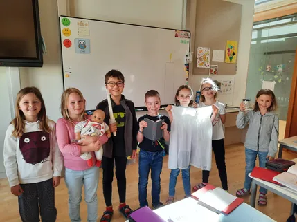 Eine Gruppe von Kindern posiert für ein Foto im Klassenzimmer, jedes Kind hält ein Objekt. Ein Mädchen hält eine Puppe, ein Junge ein Schwert und ein anderes ein Buch. Ein Mädchen hält ein weißes Kleidungsstück hoch.