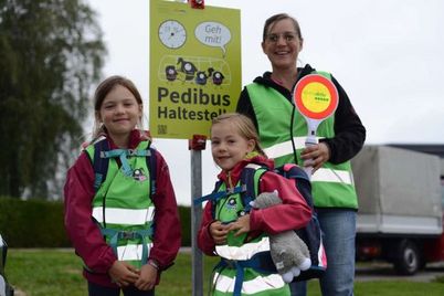Zwei junge Mädchen in Sicherheitswesten stehen neben einer Frau, die ein Schild mit der Aufschrift 'Pedibus Haltestelle' hält. Sie lächeln und scheinen auf etwas zu warten.