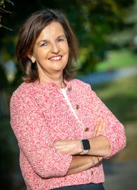 Eine Frau mit kurzen braunen Haaren, die einen rosa Pullover und ein weißes Hemd trägt, lächelt und posiert für ein Foto.