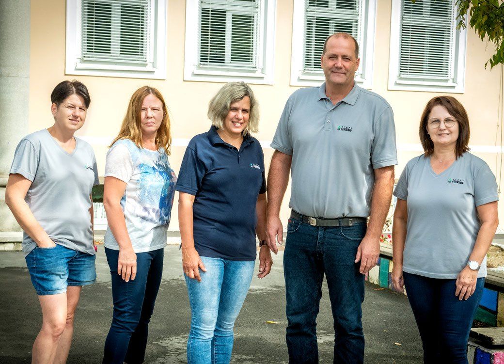 Fünf Personen stehen zusammen vor einem Gebäude. Drei Frauen und zwei Männer. Sie lächeln und scheinen für ein Foto zu posieren.