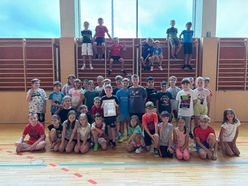 Eine Gruppe von Kindern posiert für ein Foto in einer Turnhalle. Einige Kinder sitzen auf dem Boden, während andere hinter ihnen stehen. Ein Kind vorne hält ein Zertifikat.