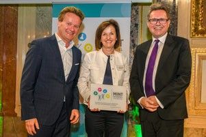 Drei Personen in formeller Kleidung stehen zusammen, zwei Männer und eine Frau, lächeln und posieren für ein Foto. Die Frau hält eine Plakette mit Symbolen und Text. Hinter ihnen ist ein Banner und ein gerahmtes Bild an der Wand.