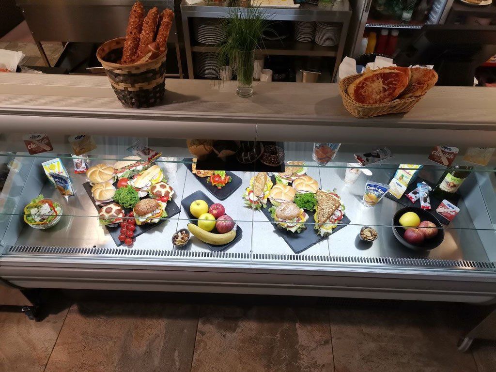 Eine Glasvitrine mit verschiedenen Lebensmitteln, darunter Sandwiches, Brot, Äpfel und Bananen, auf schwarzen Tellern angeordnet. Es gibt auch Körbe mit Brot und eine Topfpflanze auf der Theke.