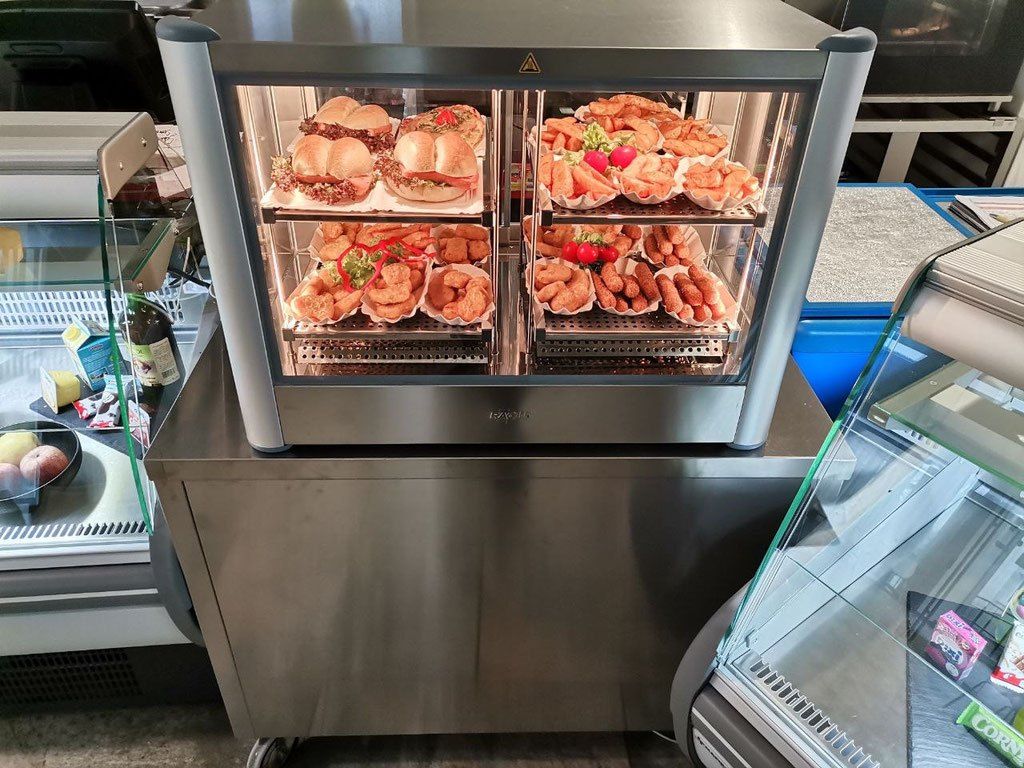 Ein Edelstahl-Lebensmitteldisplay mit drei Ebenen, auf dem verschiedene zubereitete Lebensmittel wie Sandwiches, Würstchen und Pommes Frites zu sehen sind. Der Schrank steht auf einer Metalloberfläche, mit einer Flasche in der Nähe.