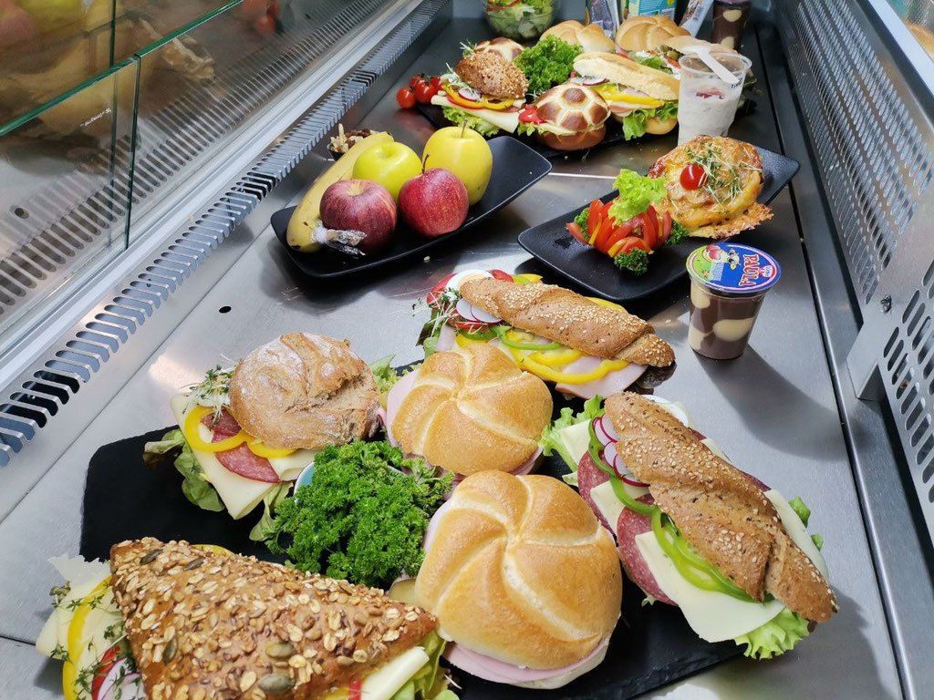Eine Auswahl an Sandwiches und Salaten, einschließlich Schinken, Käse und Gemüse, ist auf schwarzen Tellern ausgestellt. Äpfel und eine Tasse Joghurt sind ebenfalls vorhanden.