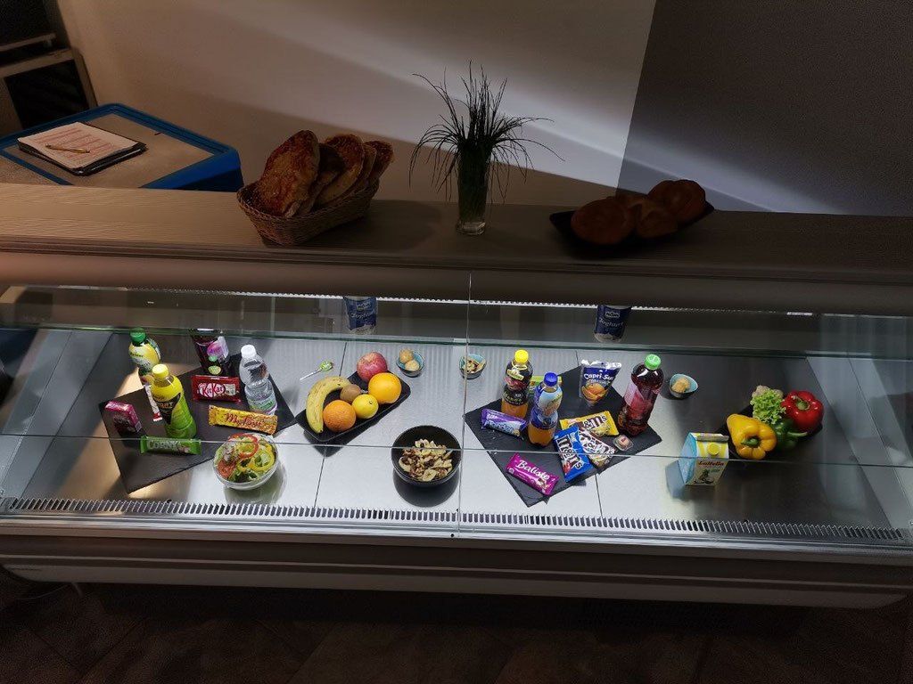 Eine Glasvitrine mit verschiedenen Lebensmitteln wie Obst, Gemüse, Müsli und Flaschen. Ein Korb mit Brot und eine Pflanze in einem Vasen stehen obenauf.