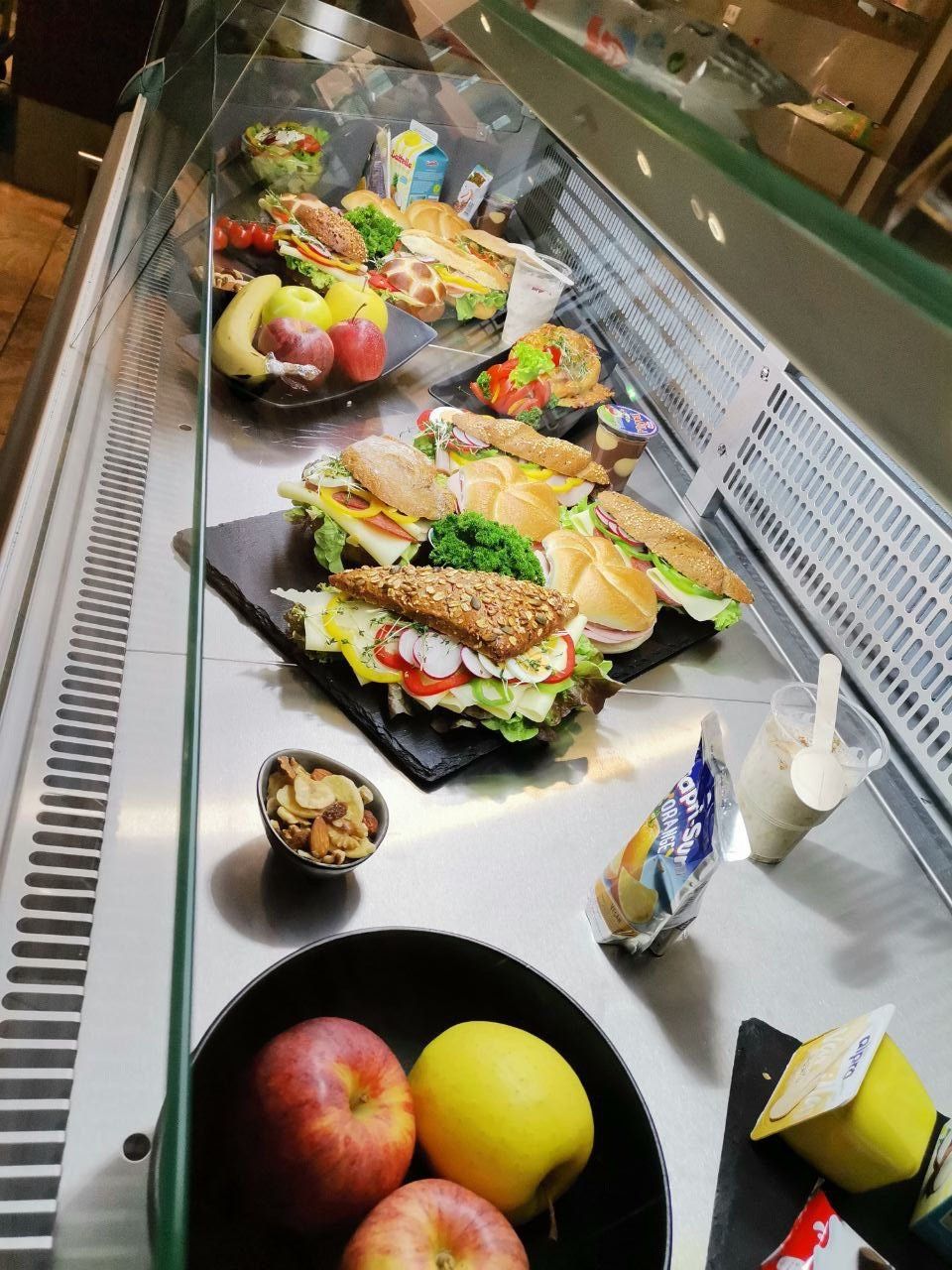 Eine Vitrine mit Sandwiches, Früchten und Getränken. Verschiedene Sandwiches, eine Banane, Äpfel und eine Schüssel mit Nüssen sind auf der linken Seite. Rechts gibt es Getränke in Bechern mit Löffeln und eine Tüte Orangensaft.