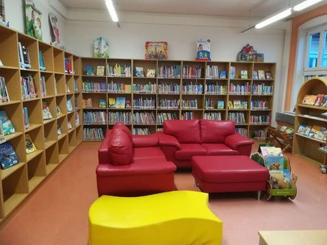 Eine Kinderbibliothek mit leuchtend roten Sofas und einem gelben Stuhl. Die Regale voller Bücher säumen die Wände, wobei Kinderbücher prominent ausgestellt sind.