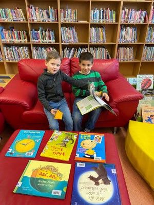 Zwei Kinder sitzen auf einer roten Couch in einer Bibliothek, umgeben von Büchern. Ein Junge liest ein Buch, während der andere ein Buch hält. Ein Tisch mit weiteren Büchern steht vor ihnen.