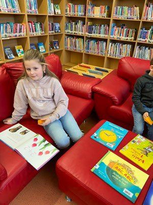 Zwei Kinder sitzen auf roten Stühlen in einer Bibliothek. Ein Mädchen hält ein Buch mit einer Blume auf dem Cover. Ein Junge sitzt neben ihr und hält ein Objekt. Bücher sind auf dem Tisch vor ihnen.