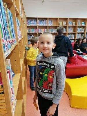 Ein junger Junge mit lächelndem Gesicht steht in einer Bibliothek und trägt eine graue Kapuzenjacke mit einem Lego-Charakter-Design. Hinter ihm befinden sich mit Büchern gefüllte Holzregale, und einige Personen sitzen auf roten und gelben Stühlen.