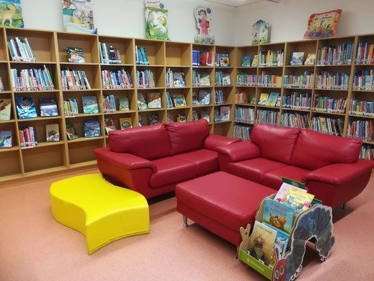 Eine Kinderbibliothek mit roten Sofas und Regalen voller bunter Bücher. Ein gelber Stuhl und ein roter Ottoman mit Büchern im Vordergrund.