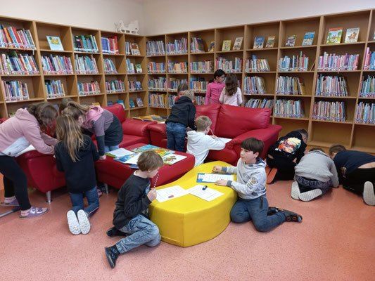 Mehrere Kinder sitzen in einer Bibliothek und studieren Bücher auf einem gelben Tisch. Sie sind von Bücherregalen umgeben, die mit Büchern gefüllt sind.