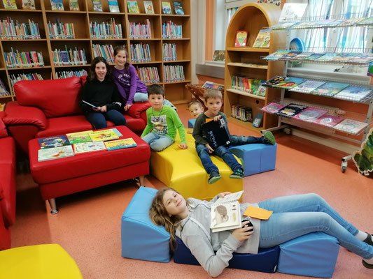 Eine Gruppe von Kindern in einer Bibliothek mit bunten Stühlen, Büchern und einer Frau, die auf einem roten Sofa sitzt.