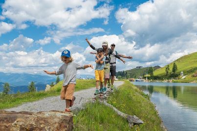 Eine fünfköpfige Familie läuft auf einem Schotterweg in der Nähe eines Sees. Die Kinder tragen Mützen und Turnschuhe, und die Erwachsenen tragen Sonnenbrillen.