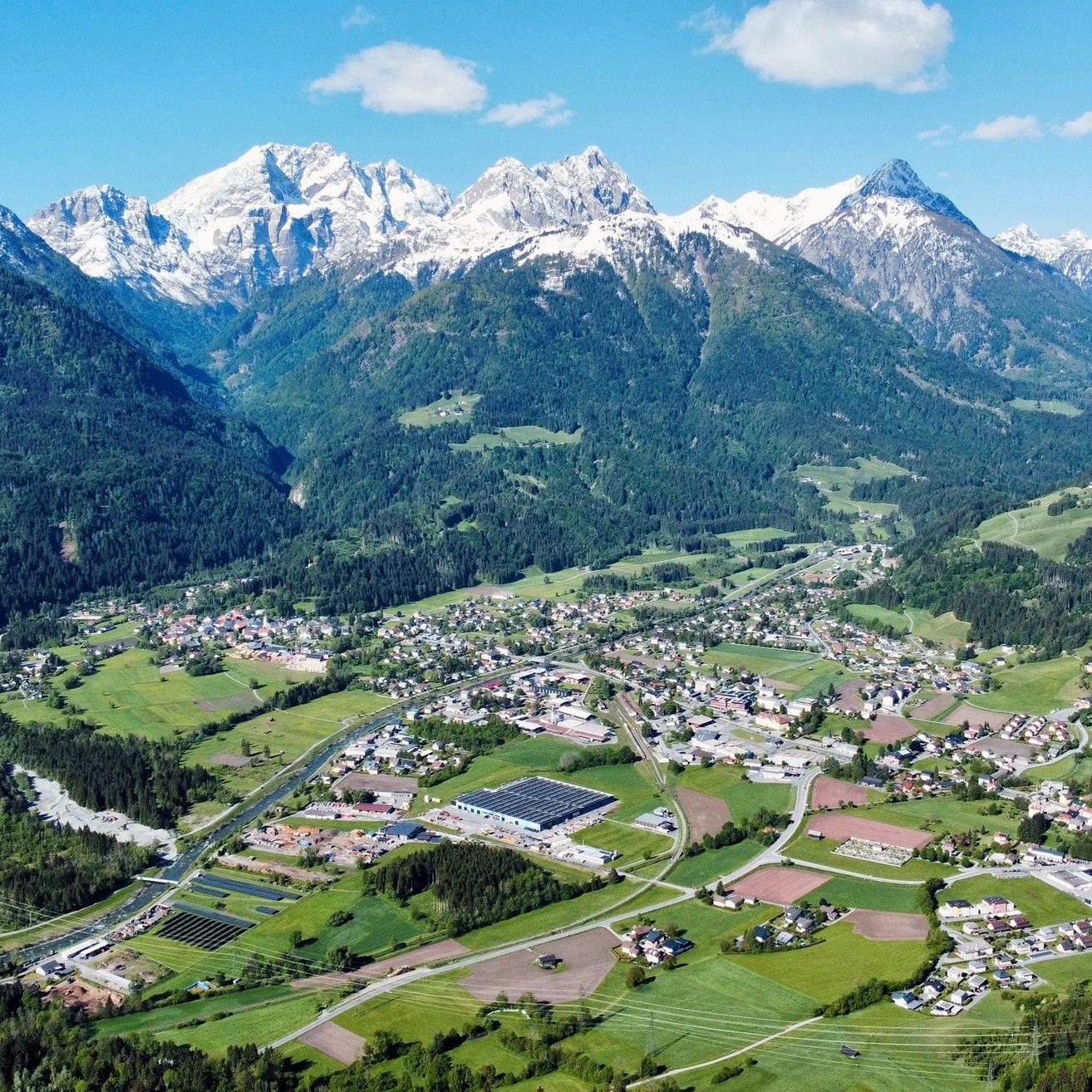 Ein Luftbild einer Bergregion mit schneebedeckten Gipfeln, einer kleinen Stadt im Tal und üppigen grünen Feldern und Bäumen.