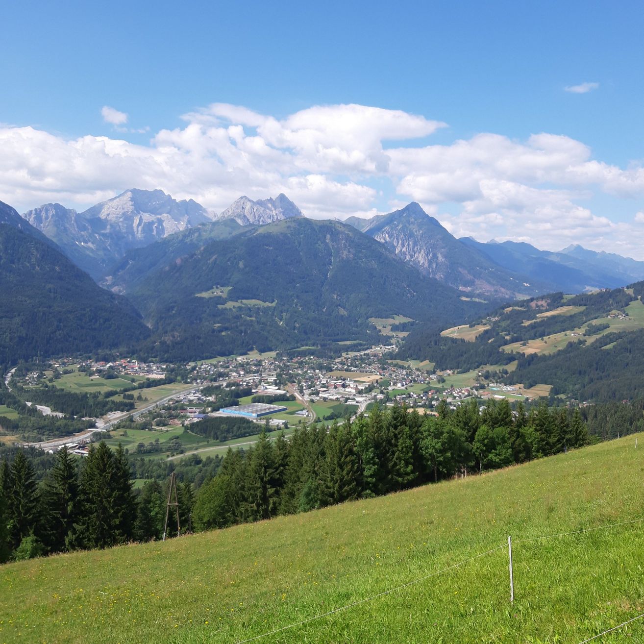 Ein Panoramablick auf ein Tal mit einer Stadt in der Mitte, umgeben von Bergen und Grün unter einem blauen Himmel mit Wolken.