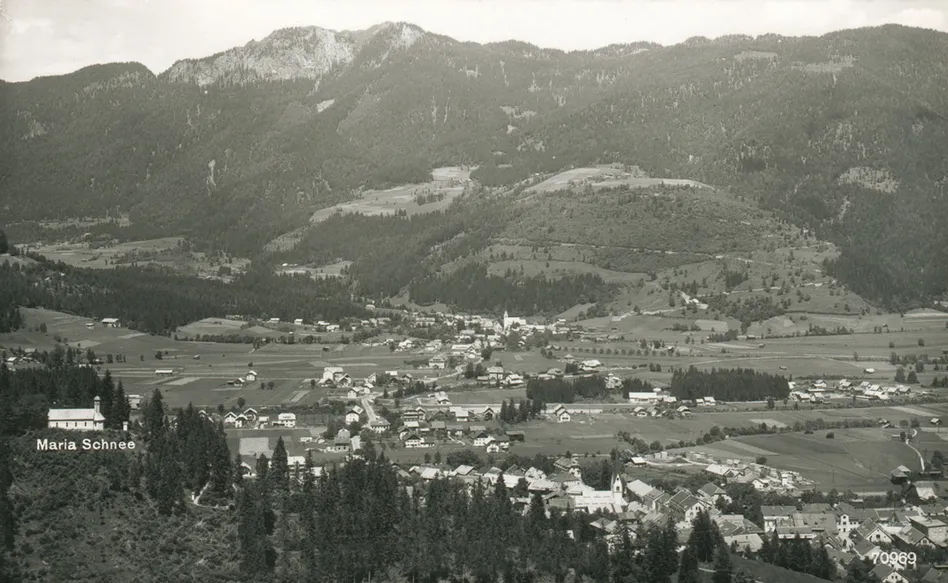 Eine Schwarz-Weiß-Luftaufnahme eines kleinen Dorfes in den Bergen, umgeben von dichten Wäldern und weitläufigen Feldern.