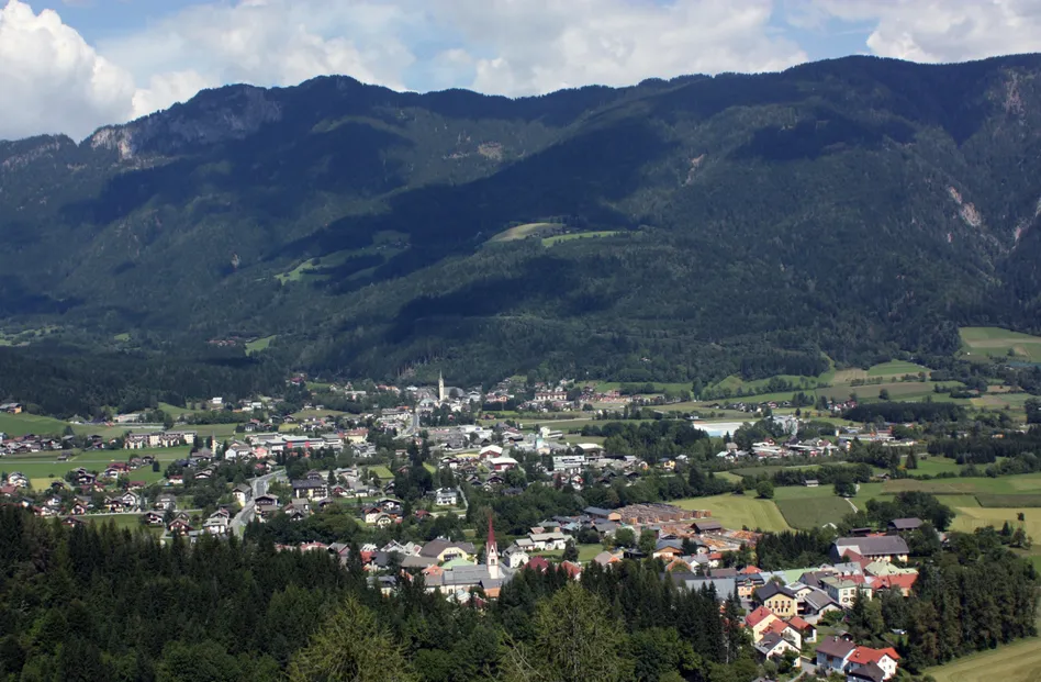 Luftaufnahme einer Stadt, umgeben von Bergen und üppiger Grünfläche, mit einem Kirchturm im Zentrum sichtbar.
