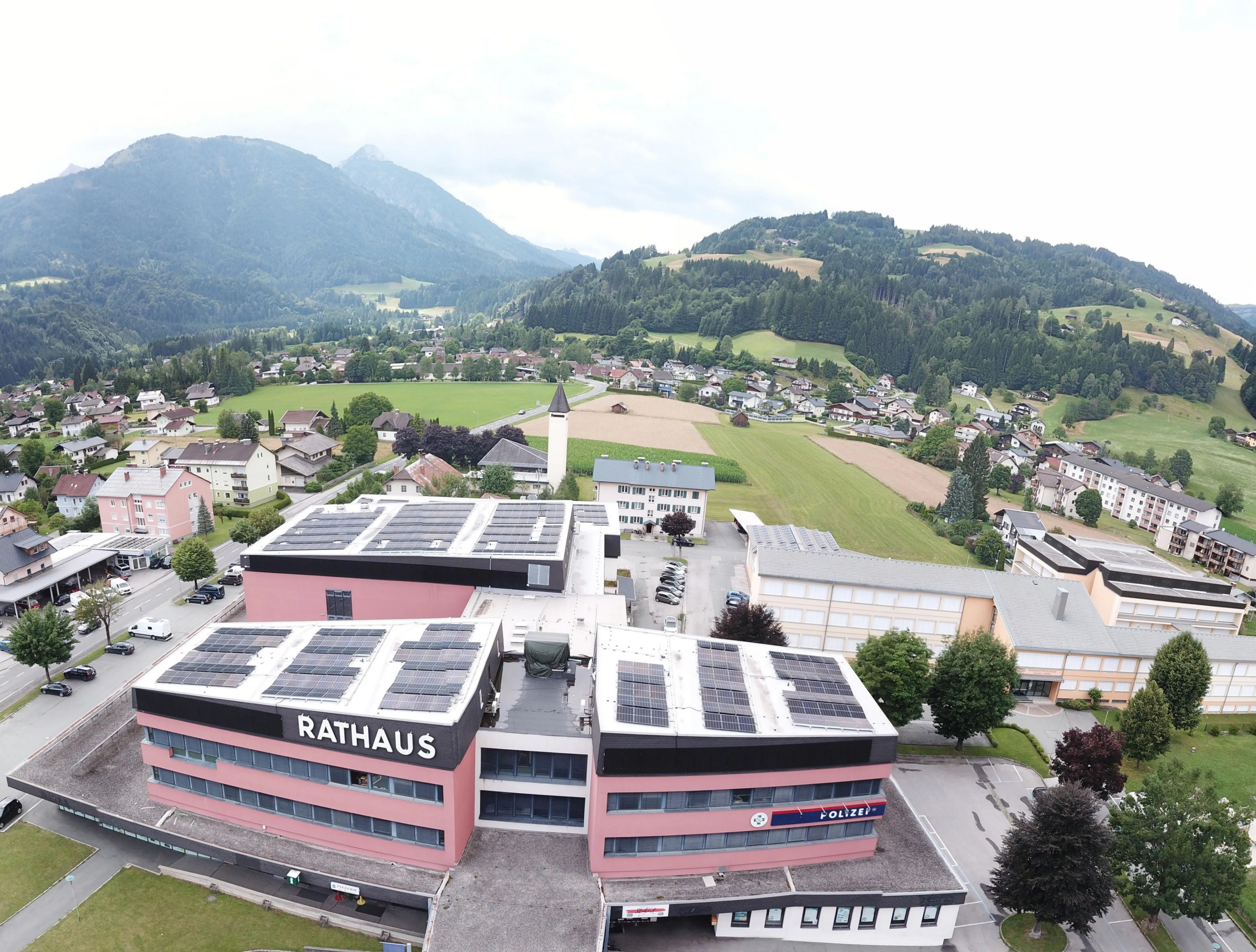 Luftaufnahme einer Stadt mit einem Rathausgebäude und einer Polizeistation, beide mit Solarpanels. Die Stadt liegt zwischen Bergen und grünen Feldern.