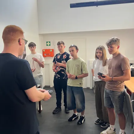 Eine Gruppe junger Menschen, möglicherweise Schüler, stehen in einem Raum. Sie alle schauen auf den Mann vor ihnen, der eine Fernbedienung in der Hand hält. Der Raum hat weiße Wände und eine weiße Decke.