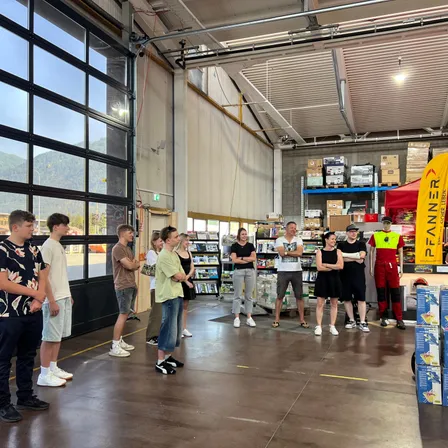 Eine Gruppe von Menschen steht in einem Lagerhaus und sieht auf ein Banner. Einige tragen Freizeitkleidung, während andere Sicherheitsausrüstung tragen. Im Hintergrund befinden sich Regale mit verschiedenen Gegenständen und ein Zelt.