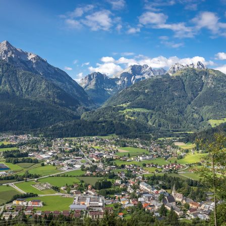 Luftaufnahme eines Dorfes, umgeben von grünen Feldern und Bergen unter einem blauen Himmel mit einigen Wolken.