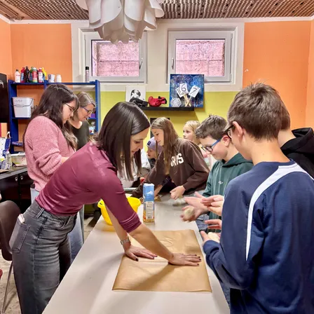 Eine Gruppe von Kindern und Erwachsenen in einem Raum, sie bereiten mit einer Frau Essen zu. Die Frau steht und bereitet etwas auf einem Tisch zu.