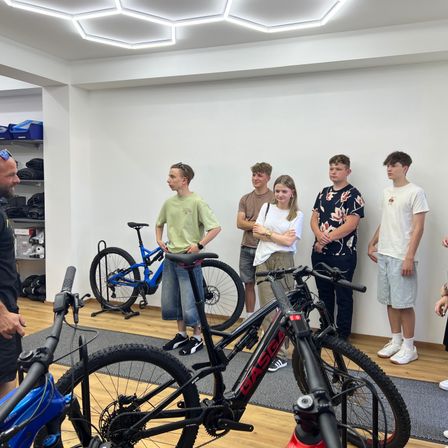 Eine Gruppe von Menschen steht in einem Raum mit Fahrrädern. Der Mann vorne erklärt ihnen etwas. Ein Fahrrad mit dem Wort Gass darauf steht auf dem Boden.