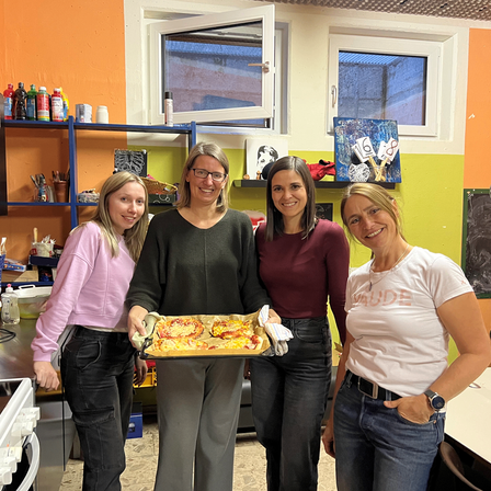 Vier Frauen stehen in einer Küche, eine hält eine Pizzaschale. Sie lächeln und scheinen für ein Foto zu posieren. Die Küche hat eine Theke, Regale und ein Fenster.