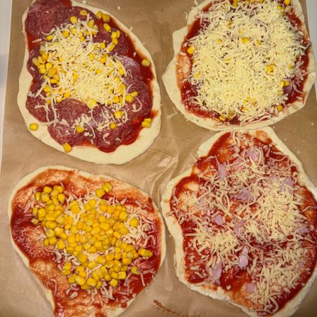 Vier selbstgemachte Pizzen liegen auf Backpapier. Jede hat rote Soße, Käse und Beläge wie Peperoni, Mais und Schinken.