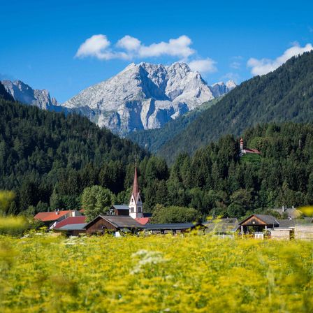 Ein malerisches Dorf mit einem Kirchturm im Vordergrund, eingebettet zwischen üppigen grünen Bäumen und Bergen unter einem blauen Himmel.