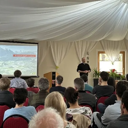 Ein Mann steht vor einer Projektionsleinwand und spricht zu einem sitzenden Publikum in einem Raum mit weißer Decke und Vorhängen.