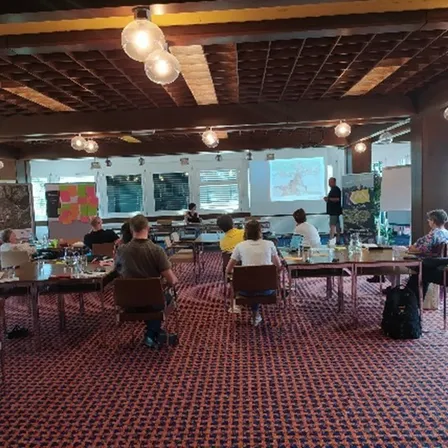 Eine Gruppe von Menschen in einem Klassenzimmer sitzt an Tischen in Stühlen. Ein Mann steht vorne im Raum vor einer Projektionsleinwand.