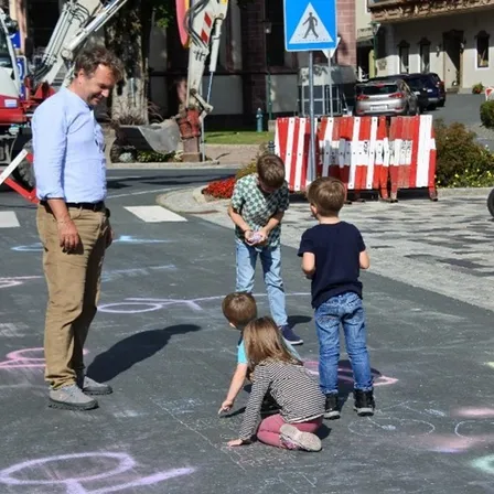 Ein Mann und vier Kinder zeichnen auf einer Straße. Ein Baufahrzeug ist in der Nähe geparkt.