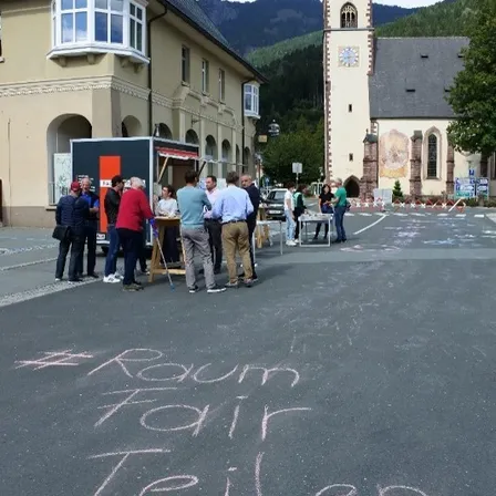 Eine Gruppe von Menschen versammelt sich vor einem Gebäude mit einer Turmspitze im Hintergrund. Ein Tisch ist aufgestellt, um den sich Menschen versammelt haben. Der Boden hat Kreideaufschriften.