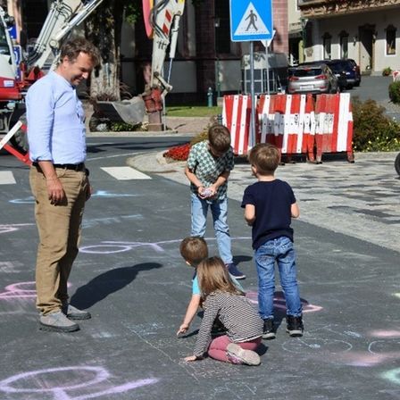 Ein Mann und vier Kinder zeichnen auf einer Straße. Ein Baufahrzeug ist in der Nähe geparkt.