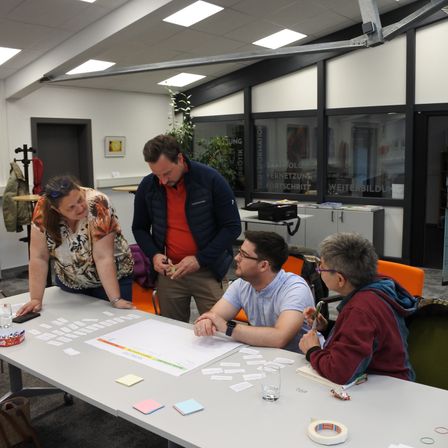 Vier Personen sind um einen Tisch versammelt und beschäftigen sich mit einer gemeinsamen Aktivität. Zwei sitzen, einer steht und einer lehnt sich über den Tisch. Verschiedene Papiere und Haftnotizen sind verteilt, und es gibt Stifte und Gläser auf dem Tisch. Der Raum hat weiße Wände und große Fenster.