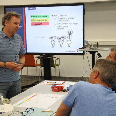 Ein Mann präsentiert bei einer Besprechung mit einem Projektionsbildschirm hinter ihm, der ein Diagramm mit Pfeilen zeigt. Zwei Männer sitzen vor ihm und hören zu. Papiere und ein Laptop liegen auf dem Tisch.