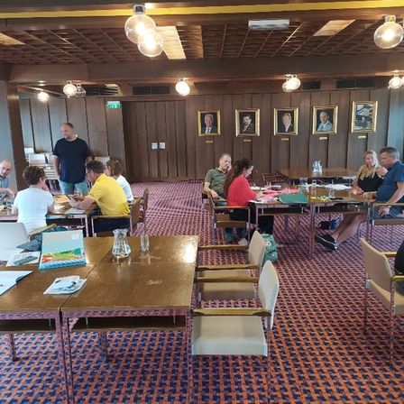 Eine Gruppe von Menschen sitzt an Tischen in einem Raum. Der Raum hat einen Teppichboden und Holzwände. An der Wand hängen Bilderrahmen. Der Raum hat mehrere Lampen, die von der Decke hängen.