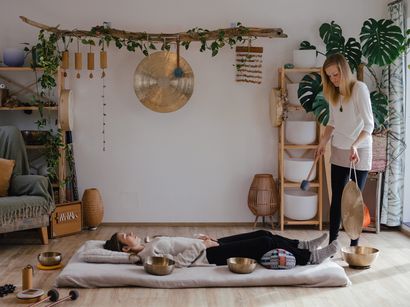 Eine Frau liegt auf einer Yogamatte, umgeben von verschiedenen Musikinstrumenten. Eine andere Frau steht in der Nähe und hält einen Hammer. Der Raum ist mit Pflanzen und dekorativen Gegenständen gefüllt.