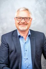 Ein Mann mit grauem Haar trägt ein blaues Hemd und einen schwarzen Anzug mit einer Brille und lächelt.