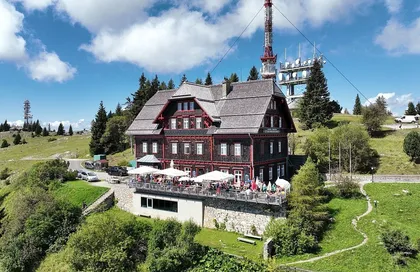 Ein Chalet-Stil Gebäude mit roten Verzierungen steht auf einem Hügel, umgeben von Bäumen. Ein Balkon mit vielen Menschen und Schirmen ist sichtbar. Ein Turm mit Antennen ist im Hintergrund.