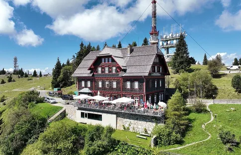 Ein Chalet mit großem Balkon und einem Turm mit Satellitenschüsseln im Hintergrund. Leute versammeln sich auf dem Balkon mit weißen Sonnenschirmen.