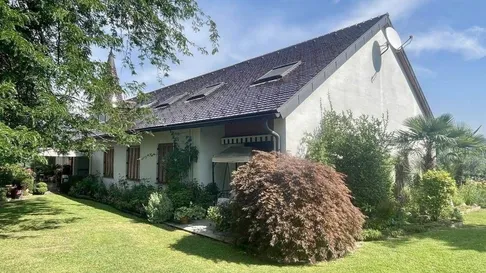 Ein weißes Haus mit einem Ziegeldach, Solarpanelen und einer Satellitenschüssel. Das Haus ist von einem Garten mit verschiedenen Pflanzen und Bäumen umgeben.