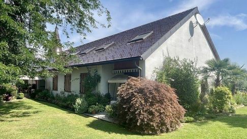 Ein weißes Haus mit einem Ziegeldach, Solarpanelen und einer Satellitenschüssel. Das Haus ist von einem Garten mit verschiedenen Pflanzen und Bäumen umgeben.
