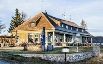 Ein großes Gebäude mit einem braunen Dach, mehreren Fenstern und einem Namensschild, das 'Alpenglückshotel' sagt. Mehrere Menschen sitzen draußen auf Stühlen. Bäume und Gras umgeben das Gebäude. Ein geschlossener blauer Regenschirm steht davor.