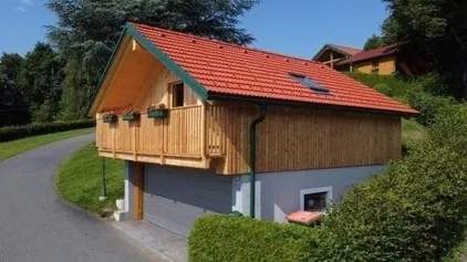 Ein kleines Holzhaus mit rotem Dach und Balkon. Es liegt an einem Hang mit einer Einfahrt und ist von Grün umgeben.